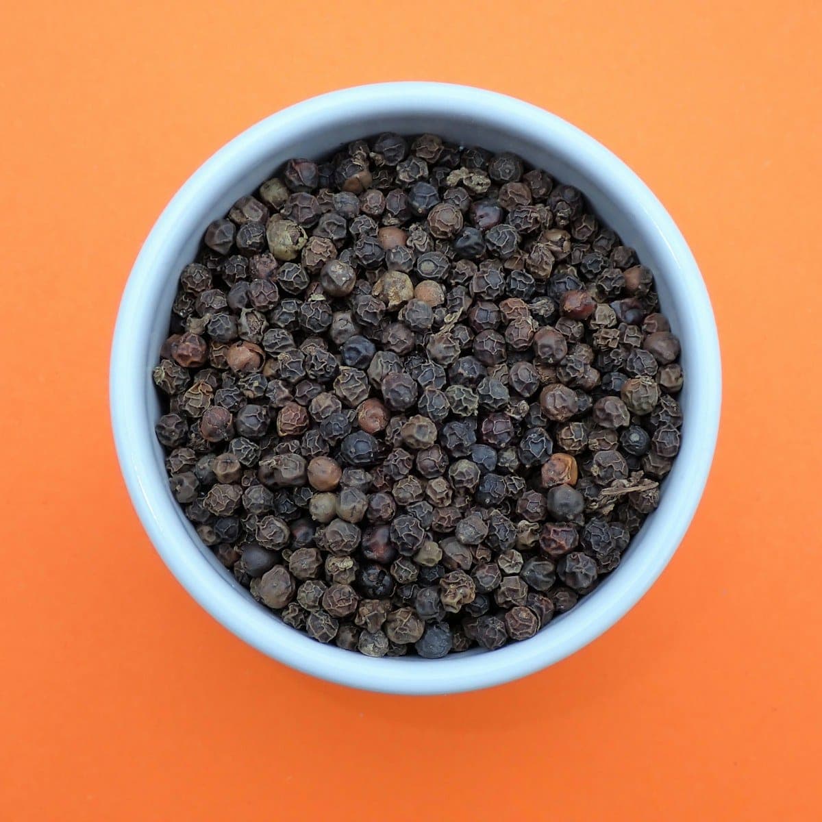Black peppercorns filling a small bowl on a warm orange ground